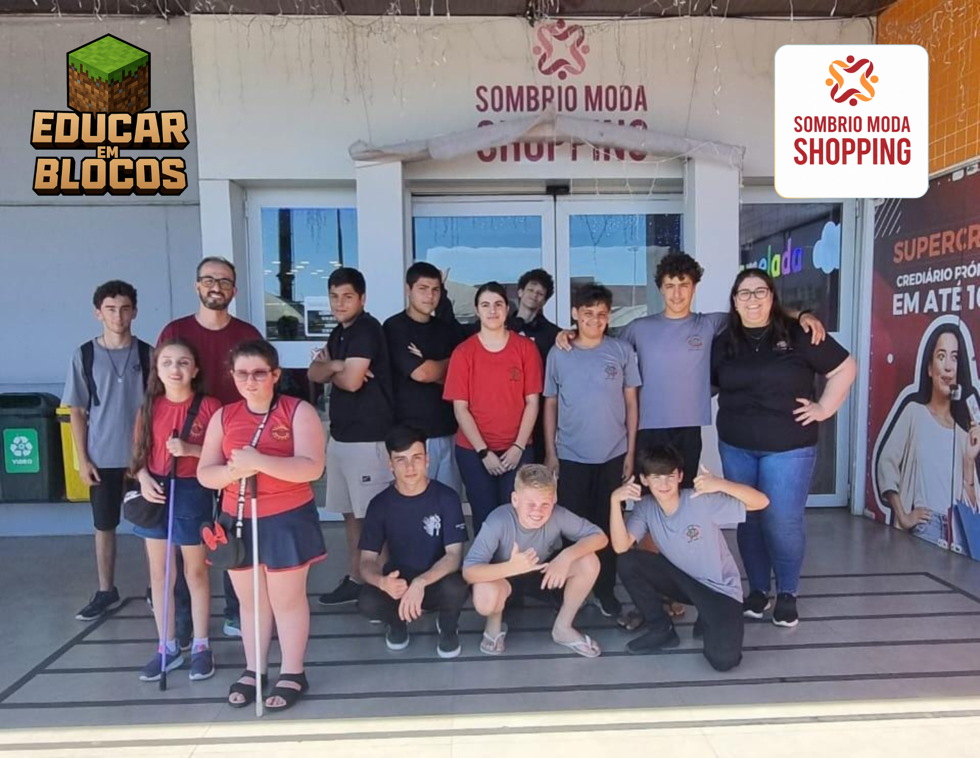 A imagem mostra um grupo de cerca de quinze pessoas reunidas em frente à entrada do Sombrio Moda Shopping. Entre elas estão jovens e alguns adultos, parte vestindo camisetas vermelhas ou cinzas, são alunos da escola Normélio Cunha e Mota Pires Na frente, duas participantes seguram bengalas brancas, indicando que são pessoas com deficiência visual, o que reforça o caráter inclusivo da atividade. O ambiente transmite a ideia de um passeio ou saída educativa em grupo. Ao fundo, é possível ver elementos do shopping, como uma lixeira de reciclagem, um cartaz promocional anunciando crédito parcelado em até 18 vezes e vitrines com cores chamativas.