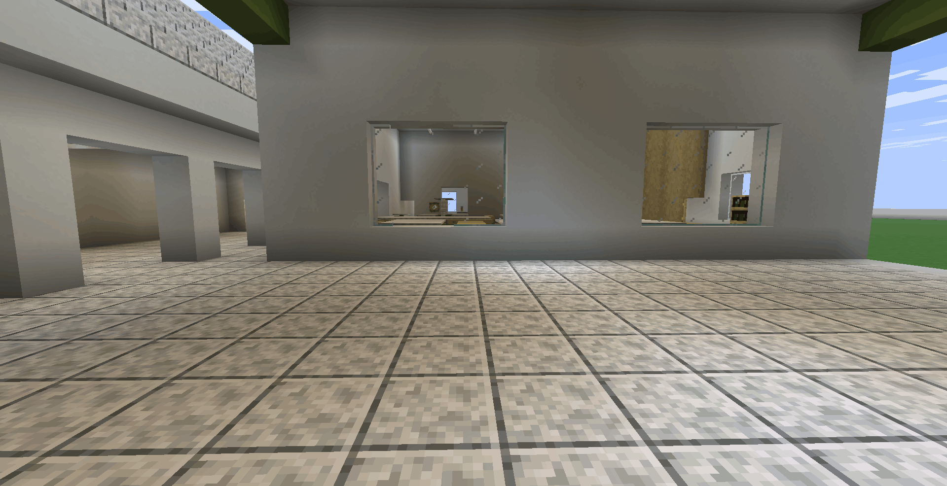 A imagem mostra a parede externa de uma sala de aula vista de dentro do pátio. O chão é todo pavimentado com pedras polidas claras. Há duas janelas de vidro quadradas que permitem ver o interior iluminado das salas.