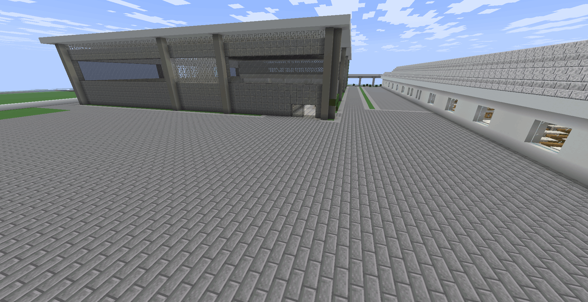 Vista ampla da área externa. À esquerda, destaca-se um grande edifício alto e robusto, feito de pedra e grades, que representa o ginásio de esportes. À direita, a lateral do prédio principal da escola. O chão entre os dois prédios é totalmente revestido com um calçamento cinza escuro