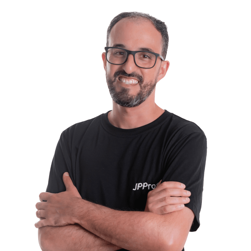 A imagem mostra um homem em pé diante de um fundo claro e liso. Ele veste uma camiseta preta com a palavra "JPPro" estampada no peito. Tem cabelo curto, barba bem aparada e usa óculos. Está sorrindo e com os braços cruzados à frente do corpo.