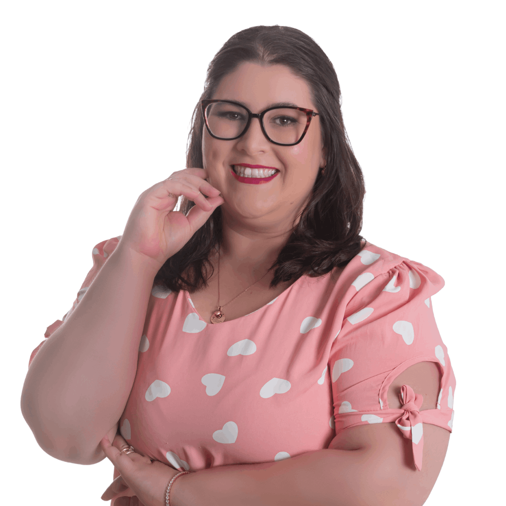 A imagem mostra uma mulher em pé diante de um fundo claro e uniforme. Ela veste uma blusa rosa com estampa de corações brancos e mangas curtas, sendo que uma das mangas tem um detalhe de laço amarrado. Usa também calça jeans azul. A mulher tem cabelo escuro na altura dos ombros, está sorrindo e usa óculos, colar, pulseira e anel. Sua pose é descontraída e confiante: uma mão toca o rosto enquanto o outro braço está cruzado.
